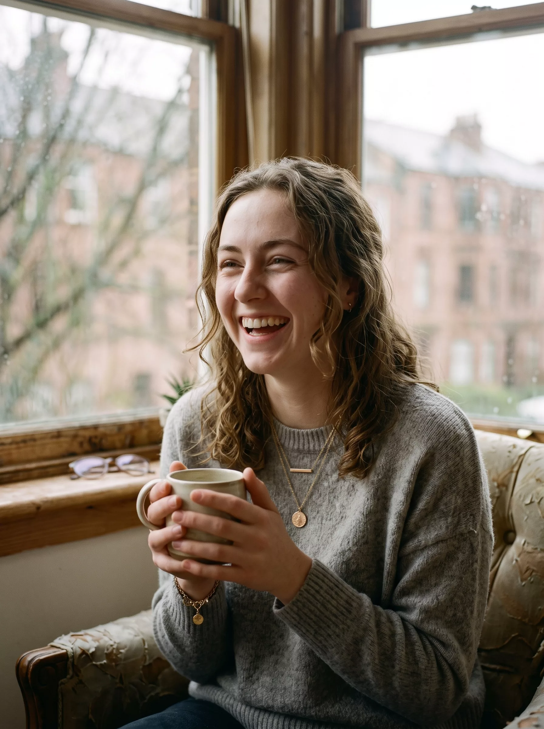 Woman laughing over a cup of coffee, wearing gold necklaces and a bracelet